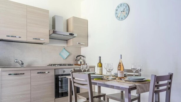 A modern kitchen with wooden cabinets, a built-in oven, and a dining table set for four with wine glasses and a bottle. - Casa Cinzia - Apartment (Vico Equense)