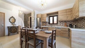 A modern kitchen with wooden cabinets, a dining table with chairs, a chandelier, and a stone wall. - Asfendamos Villas - With 2 Private Pools (Mylopotamos)