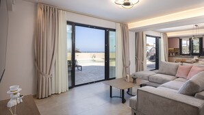 A modern living room with a large sofa, a coffee table, and a view of a pool and outdoor area. - Villa Azure Luxury Sea View Villa (Mylopotamos)