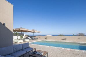 A poolside area with a sofa, lounge chairs, and umbrellas. - Villa Azure Luxury Sea View Villa (Mylopotamos)