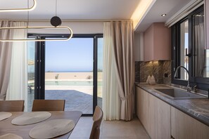 A modern kitchen with a large window, a dining area with a table and chairs, and a view of the sea. - Villa Azure Luxury Sea View Villa (Mylopotamos)