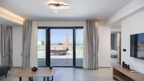 A modern hotel room with a large sliding glass door leading to a balcony, a flat-screen TV, a wooden table, and a sofa. - Villa Selene Luxury Sea View Villa (Mylopotamos)