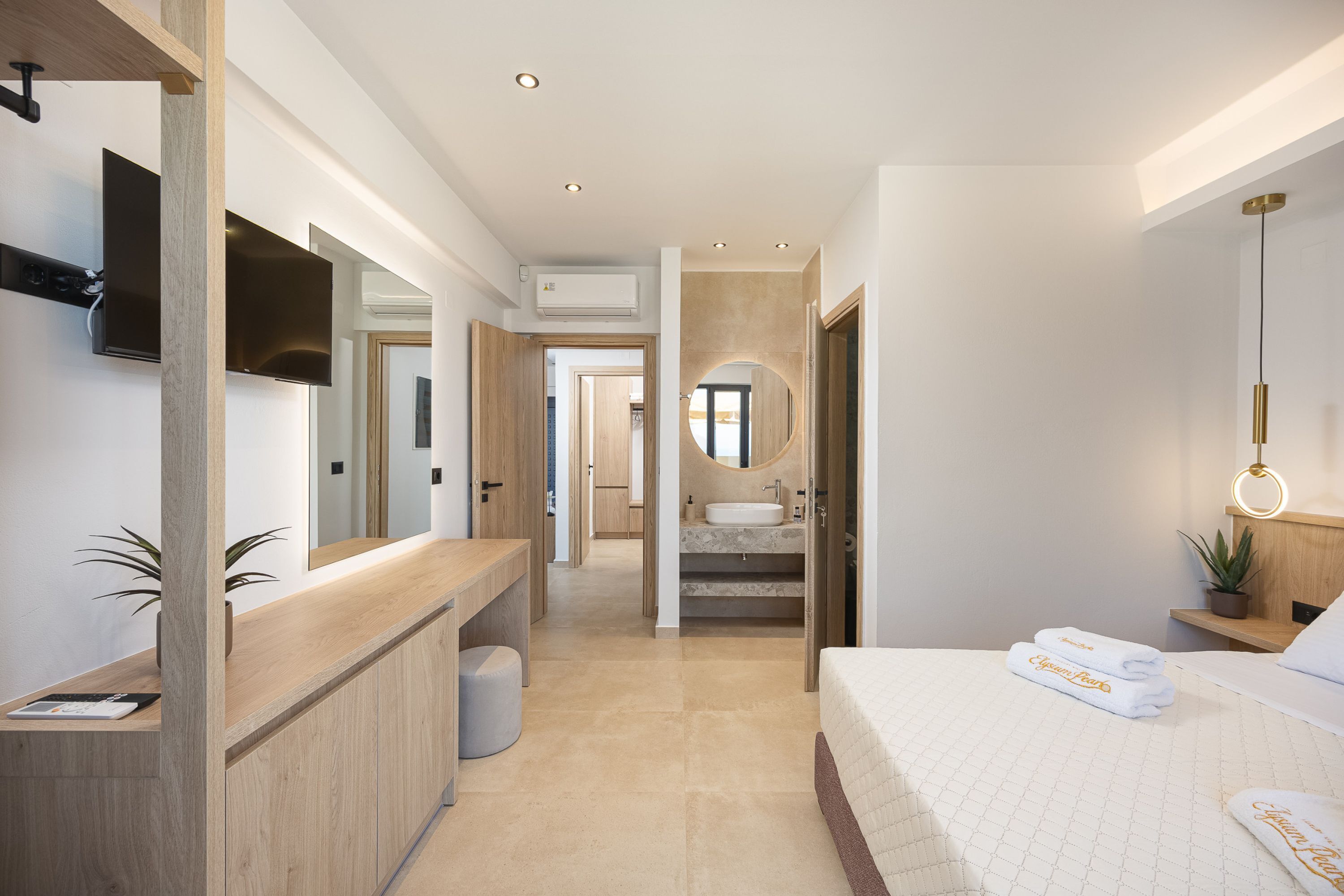 A modern hotel room with a wooden reception desk, a large bed with white linens, a round mirror, and a TV mounted on the wall.