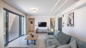 A modern living room with a sofa, a coffee table, a flat-screen TV, and a staircase leading to another level. - Elysioum Pearl Villas - With 2 Private Pools (Mylopotamos)