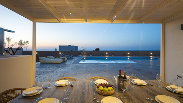 An outdoor dining area with a view of the sea, a pool, and lounge chairs. - Elysioum Pearl Villas - With 2 Private Pools (Mylopotamos)