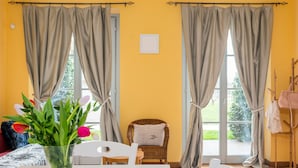 Een kamer met een gele wand, een rieten stoel, een witte tafel en een raam met gordijnen.