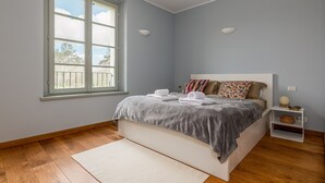 Een slaapkamer met een groot raam, een bed met kussens en handdoeken, een nachtkastje en een bijzettafeltje.
