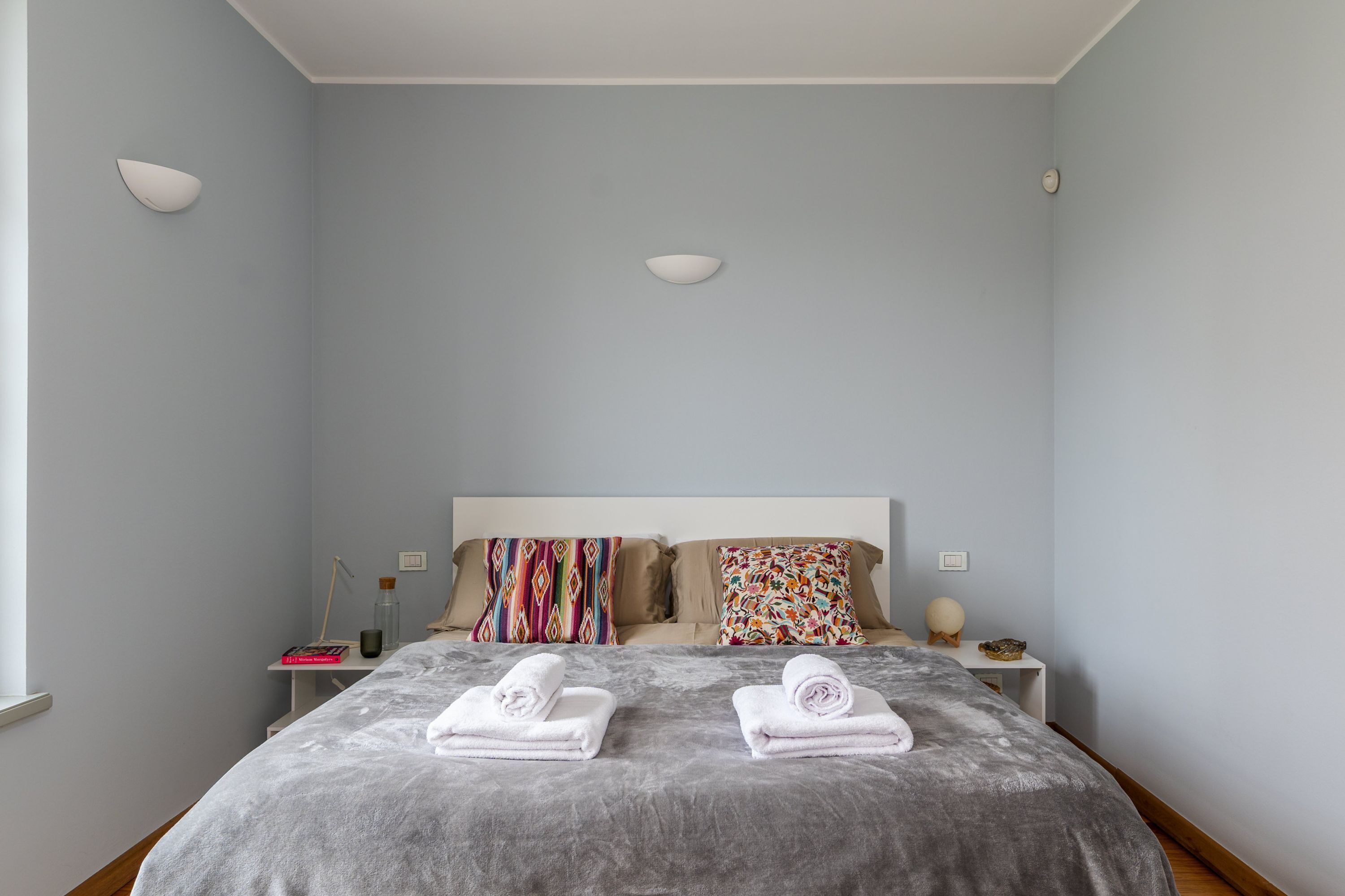 A bedroom with a bed, two bedside tables, a nightstand, a window, and a wall-mounted light fixture.