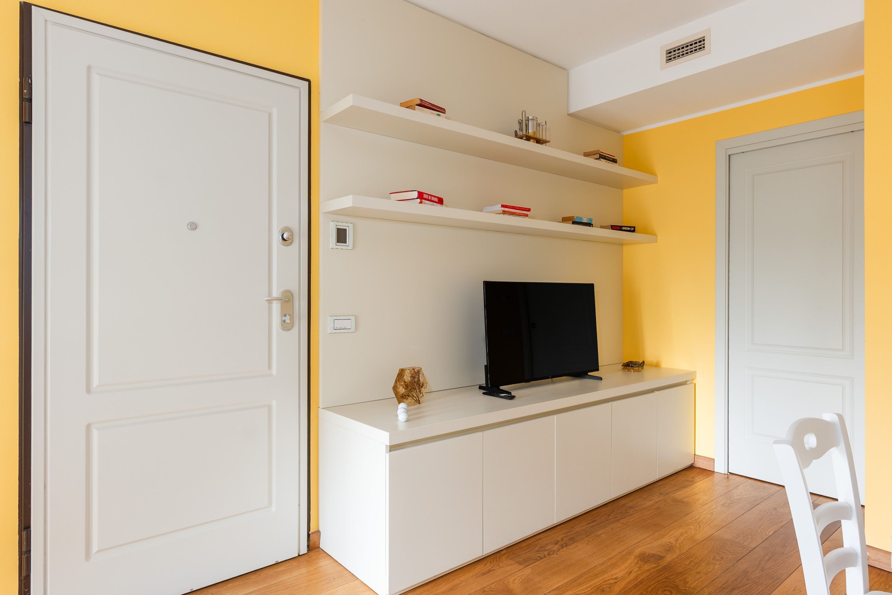 Una habitación con una puerta blanca, una consola blanca con un televisor de pantalla plana, estanterías con libros y objetos decorativos, y suelo de madera.