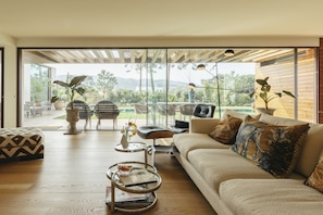 A modern living room with a sofa, coffee table, and chairs, a large window with a view of greenery and a patio. - Moon Forest Retreat by Cadenzalux (Sintra)