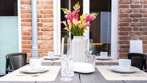 Tavola imbandita per due, con un vaso di fiori, bicchieri trasparenti e piatti e tazze bianche.