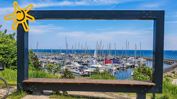 Una marina con numerose imbarcazioni ormeggiate, cielo azzurro e una panchina in legno sullo sfondo.