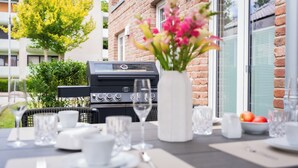 Zona pranzo all'aperto con barbecue, tavolo apparecchiato con piatti, bicchieri e un vaso di fiori.