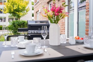 Ein Essbereich im Freien mit Grill, Tisch gedeckt mit Tellern, Gläsern und einer Blumenvase.