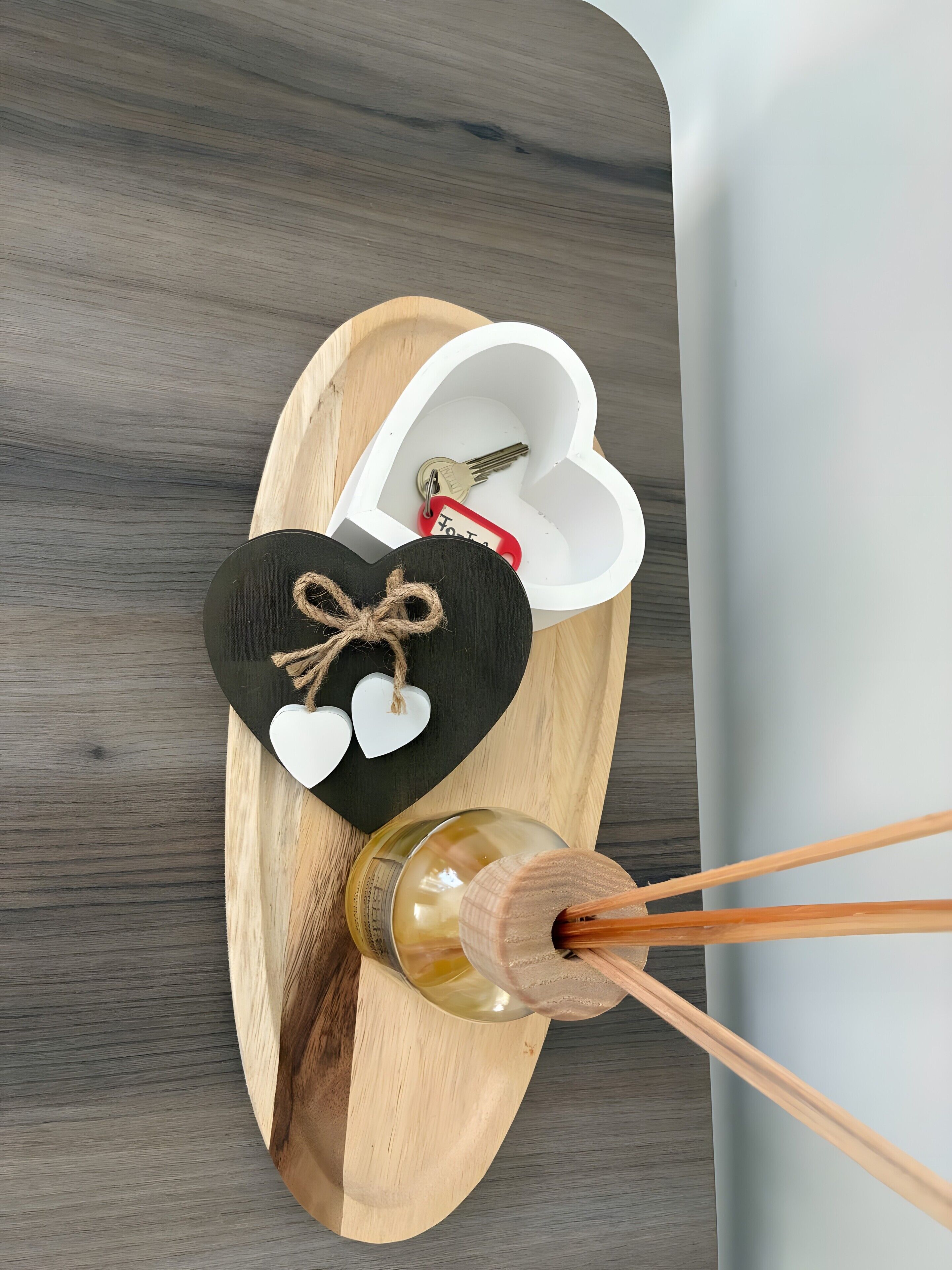Una bandeja de madera con una caja en forma de corazón que contiene llaves y una botella con un agitador de madera.