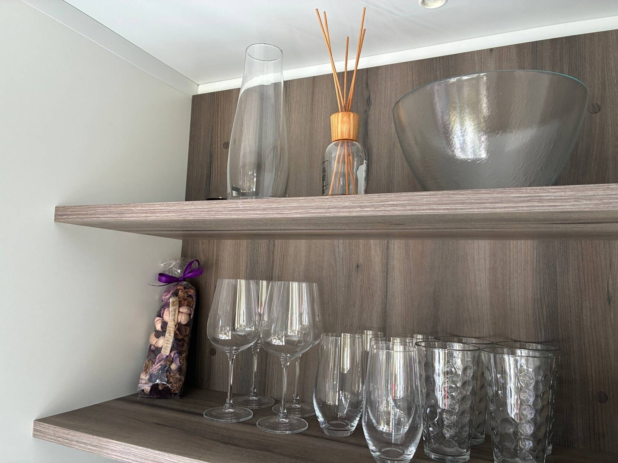 Een houten plank met een glazen vaas, rietverspreider en een schaal. Daaronder staan heldere wijnglazen en een decoratief object.