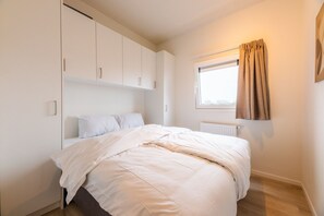 A bedroom with a bed, white cabinets, a window with curtains, and a radiator. - Enjoy stylish comfort in the Duinrand Lodge for 6 persons. This cozy and spacious holiday home offers everything you need for a truly relaxing holiday. (De Haan)