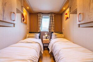 A bedroom with two beds, a nightstand, a window with curtains, and wall-mounted light fixtures. - Experience the Strandrust Lodge at Holiday Park Blankenberger Duinen at any time of the year. Enjoy a carefree holiday with the whole family. (De Haan)