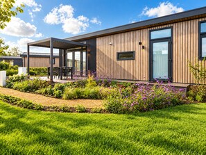 Ein modernes Gebäude mit einer überdachten Außenbestuhlung, einem gepflegten Garten mit violetten Blumen und einem klaren, blauen Himmel.