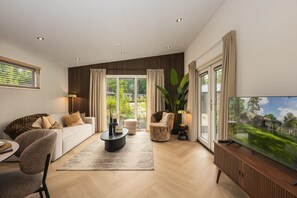 Ein modernes Wohnzimmer mit Sofa, Sesseln, Couchtisch, Fernseher und einem großen Fenster mit Vorhängen.