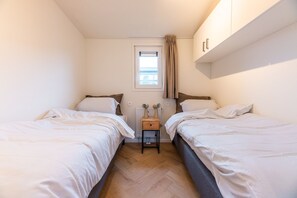 Et lille, godt oplyst rum med to senge, et vindue med gardiner, en radiator og et lille bord med en lampe.