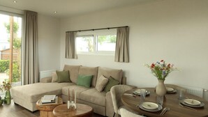 A living room with a sofa, a dining table set for two, and a window with curtains. - In the Duinrand Lodge for 4 persons, you can truly relax. This comfortable and tastefully furnished holiday home guarantees pure holiday enjoyment. (De Haan)