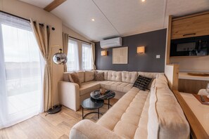 A modern living room with a sectional sofa, a small coffee table, and a dining area with a microwave. - Enjoy your holiday in the Zeebries Cabin: a comfortable holiday home on the Flemish coast for 4 persons, fully equipped for a carefree stay. (De Haan)