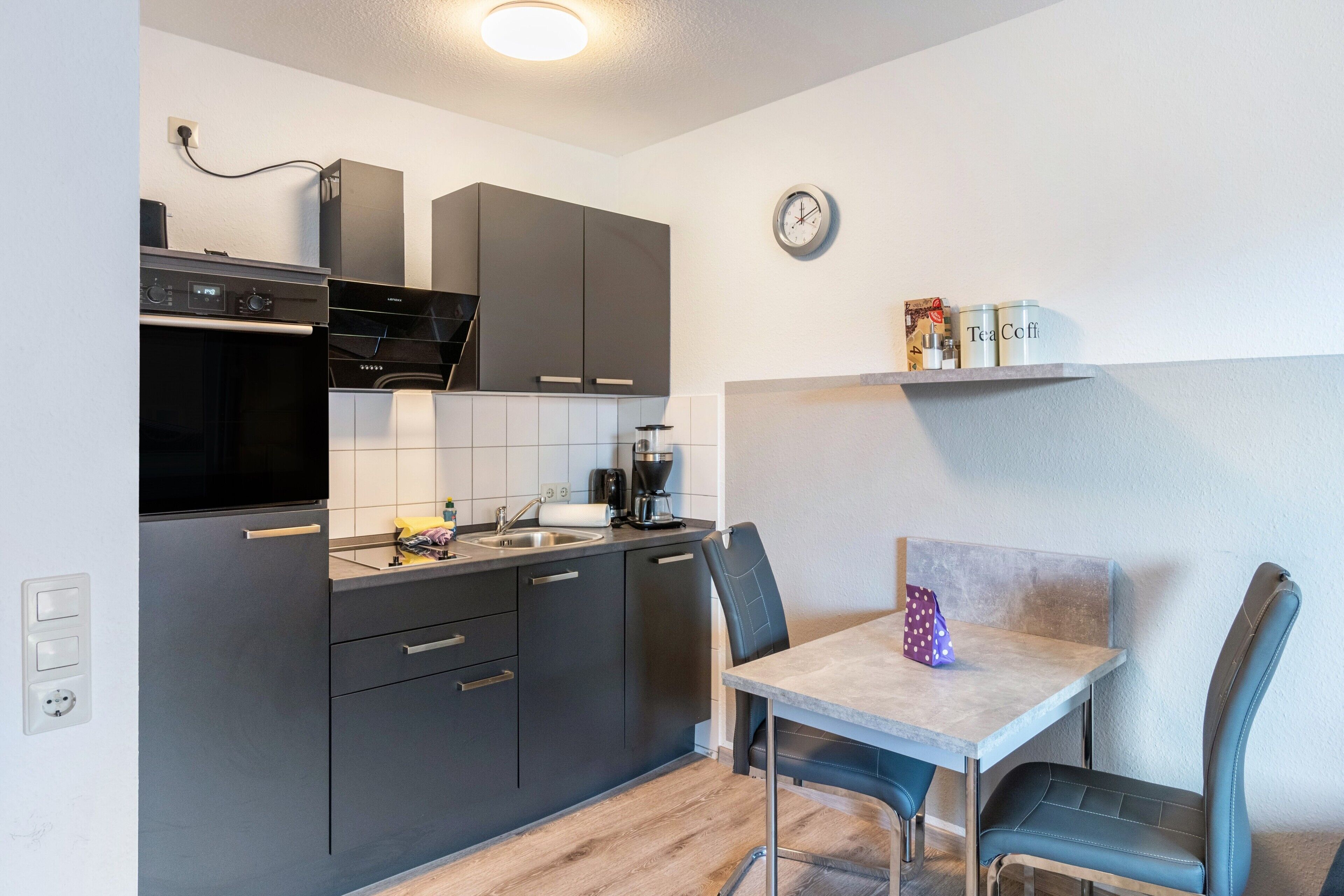 Eine kompakte Küche mit modernen Geräten, ein Essbereich mit Tisch und Stühlen und eine Uhr an der Wand.