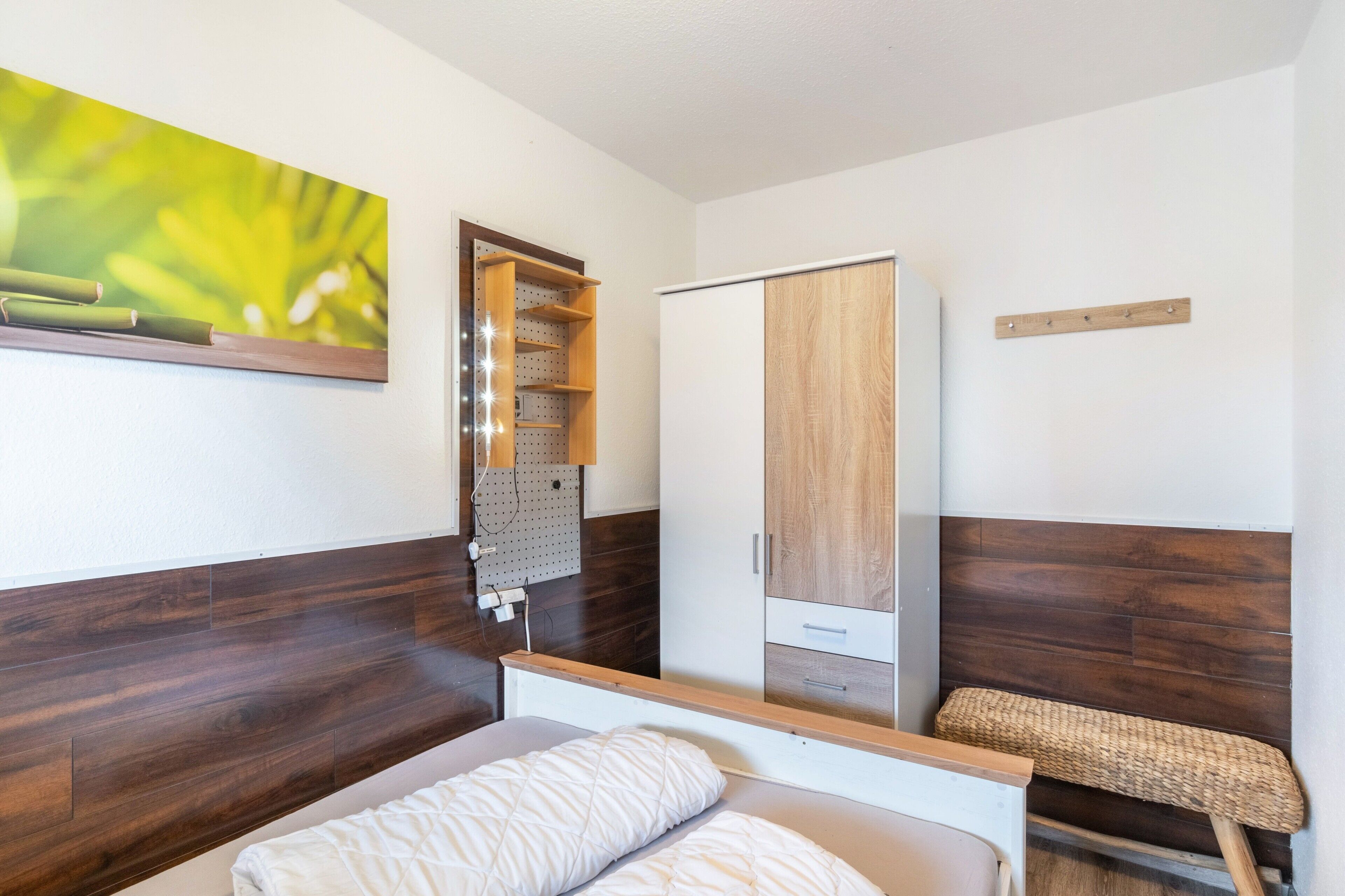 Un dormitorio con cama, armario, tablero de anuncios con estantes y una silla de mimbre.