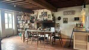 Quarto rústico com vigas de madeira aparentes, lareira, estante de livros, mesa de jantar com cadeiras e área de cozinha com micro-ondas e prateleiras.