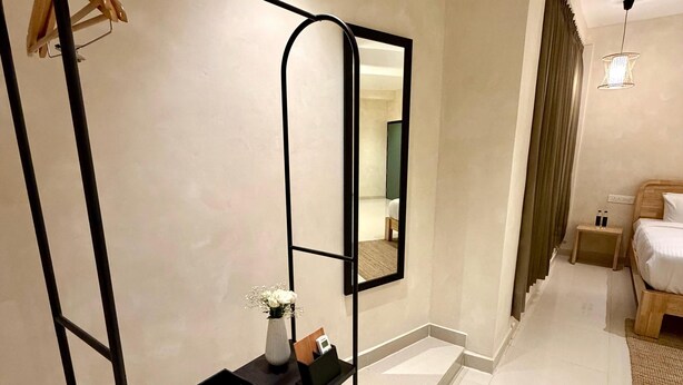Habitación de hotel con un espejo grande, un perchero metálico negro, una mesita con un florero, una cama y una mesita de noche con botellas.