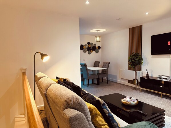 A modern living room with a sofa, a coffee table, a dining area with chairs, a TV, and a decorative wall piece. - Modern2 Bedroom Apartment/private Parking /sleep 5/balcony View in Birmingham City Centre (Birmingham)
