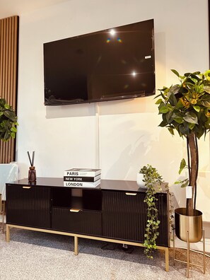 A modern living room with a flat-screen TV mounted on the wall, a black entertainment unit with books, and two potted plants. - Modern2 Bedroom Apartment/private Parking /sleep 5/balcony View in Birmingham City Centre (Birmingham)