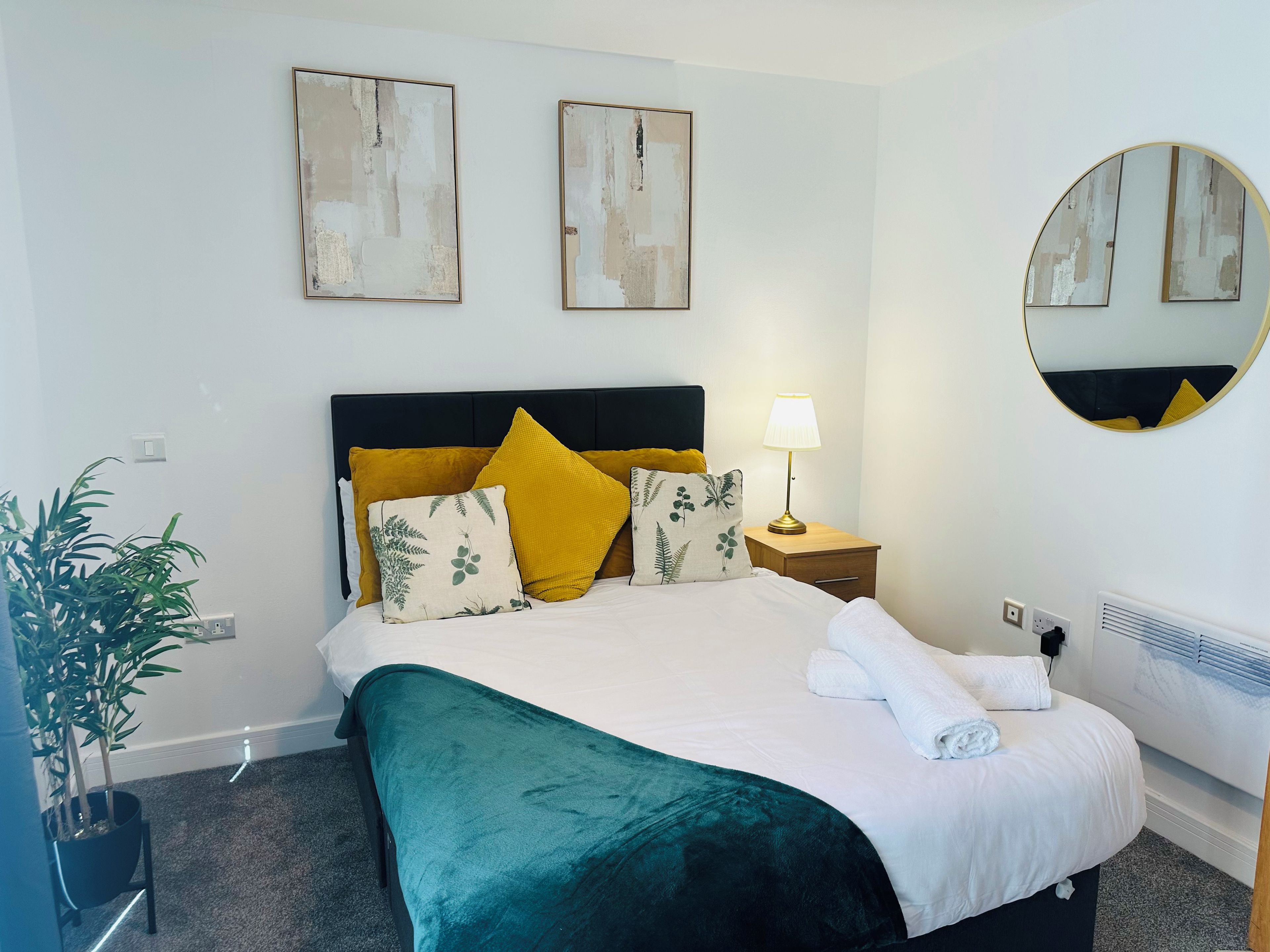 A neatly made bed with a green bedspread and multiple pillows, a bedside table with a lamp, a round mirror on the wall, and a potted plant.