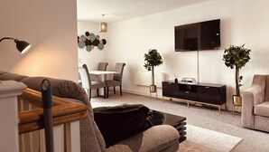Un salon moderne comprenant un canapé, une table basse, un coin repas avec des chaises, un téléviseur fixé au mur et deux plantes en pot.