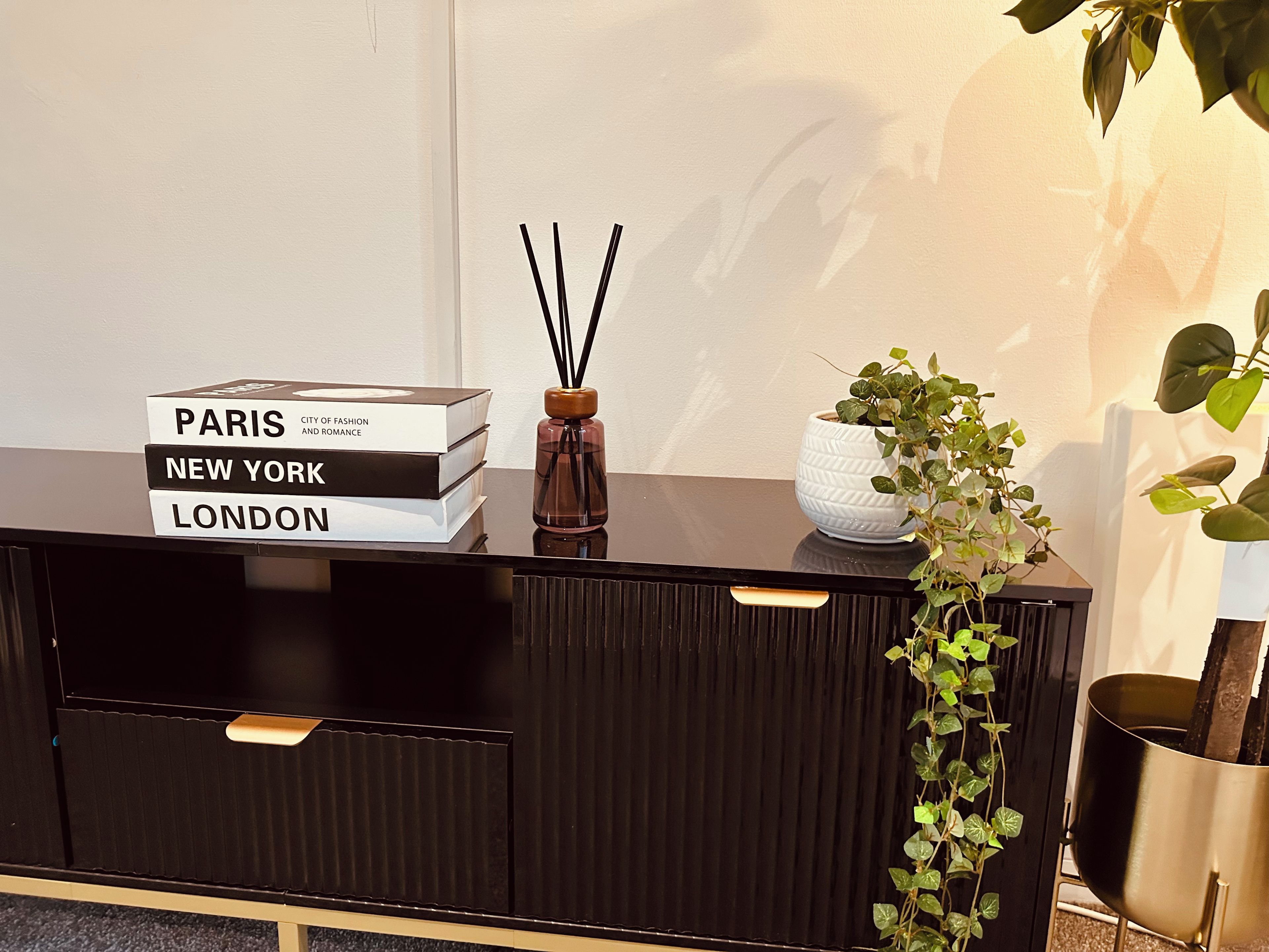 A black console table with a stack of books titled 'PARIS,' 'NEW YORK,' and 'LONDON,' a clear glass bottle with black sticks, a white pot with green plants, and a potted plant with broad leaves.