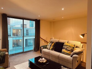 A living room with a sofa, a coffee table, and a view of a building through large windows. - Modern2 Bedroom Apartment/private Parking /sleep 5/balcony View in Birmingham City Centre (Birmingham)