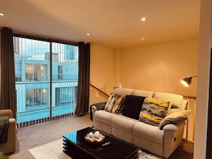 A modern living room with a sofa, a coffee table with decorative items, a lamp, and a view of a building through large windows.