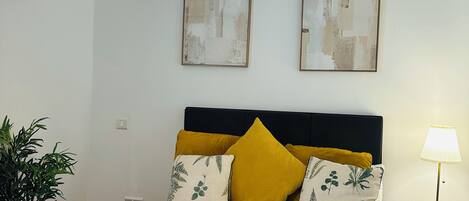Ein ordentlich bezogenes Bett mit weißen Leinen, einer türkisfarbenen Tagesdecke und zwei aufgerollten weißen Handtüchern. Ein Nachttisch mit Lampe, einer Topfpflanze und einem Wandbild sind ebenfalls zu sehen.