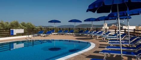 Une piscine entourée de chaises longues bleues et de parasols, le tout délimité par une clôture en bois, avec des montagnes en toile de fond.
