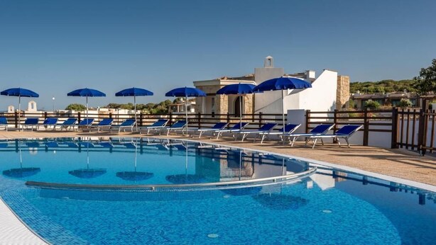 Una piscina con sillones de descanso y sombrillas, un edificio blanco de fachada de piedra y un cielo azul despejado.