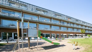 Un immeuble à plusieurs étages doté de balcons, d’un espace extérieur aménagé pour s’asseoir et d’un terrain de jeux avec des balançoires et un toboggan.