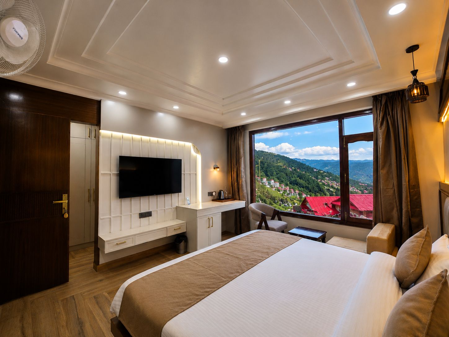 A hotel room with a large bed, a TV mounted on the wall, a desk with a chair, and a window with a view of a mountainous landscape.