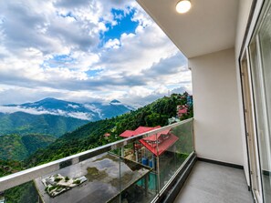 A balcony with a glass railing overlooking a mountainous landscape with houses. - The Heaven Paradise Premium (Shimla)