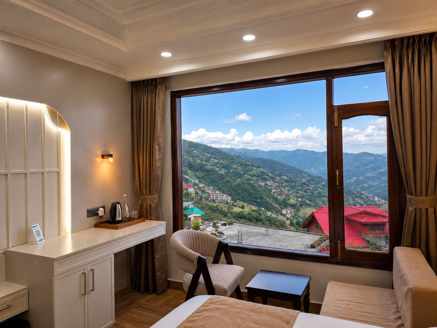 A hotel room with a bed, a desk, a chair, a small table, and a large window offering a mountain view.