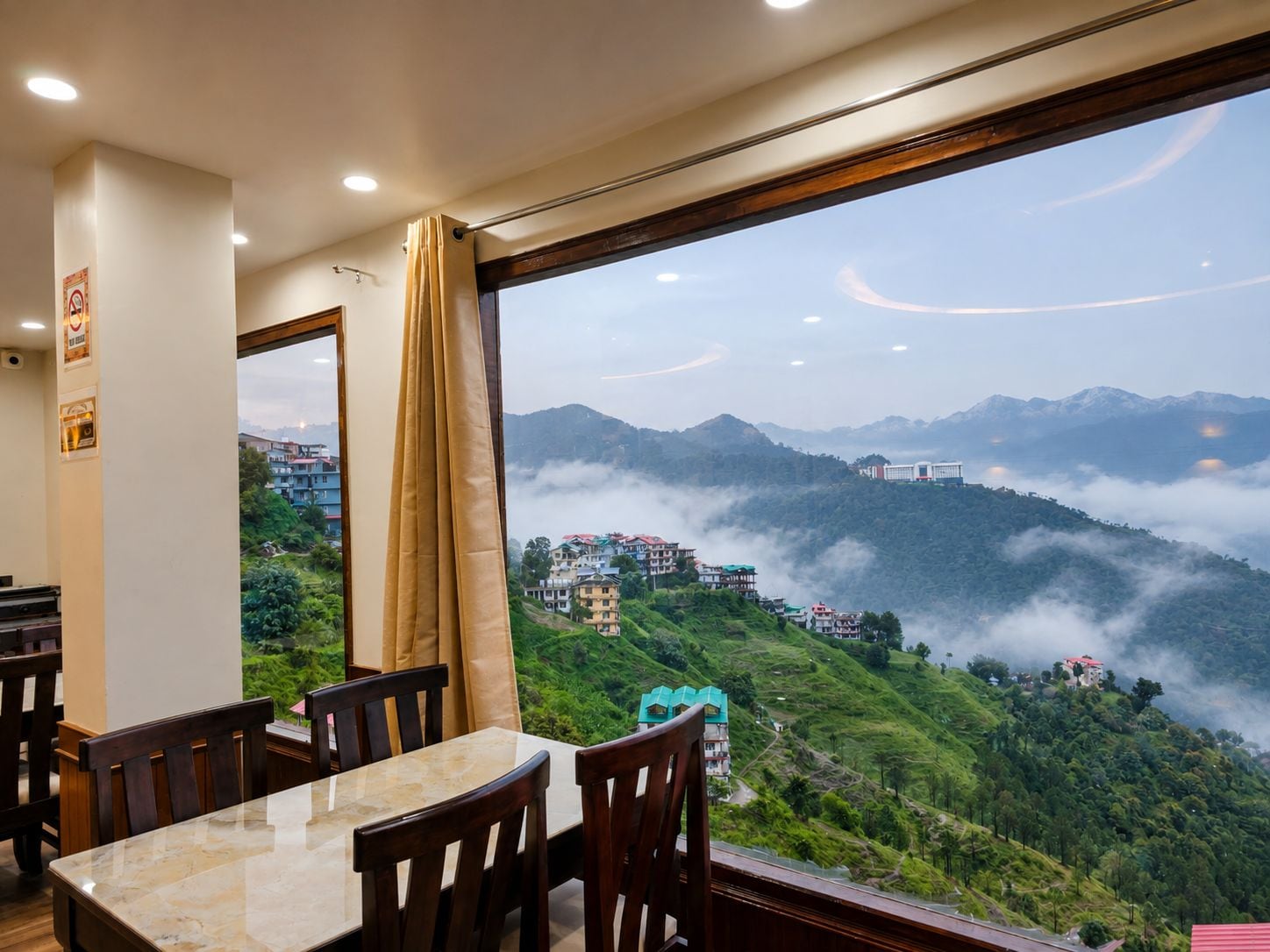 Uma sala de jantar com mesa e cadeiras, janela com cortinas, e vista para uma paisagem montanhosa.