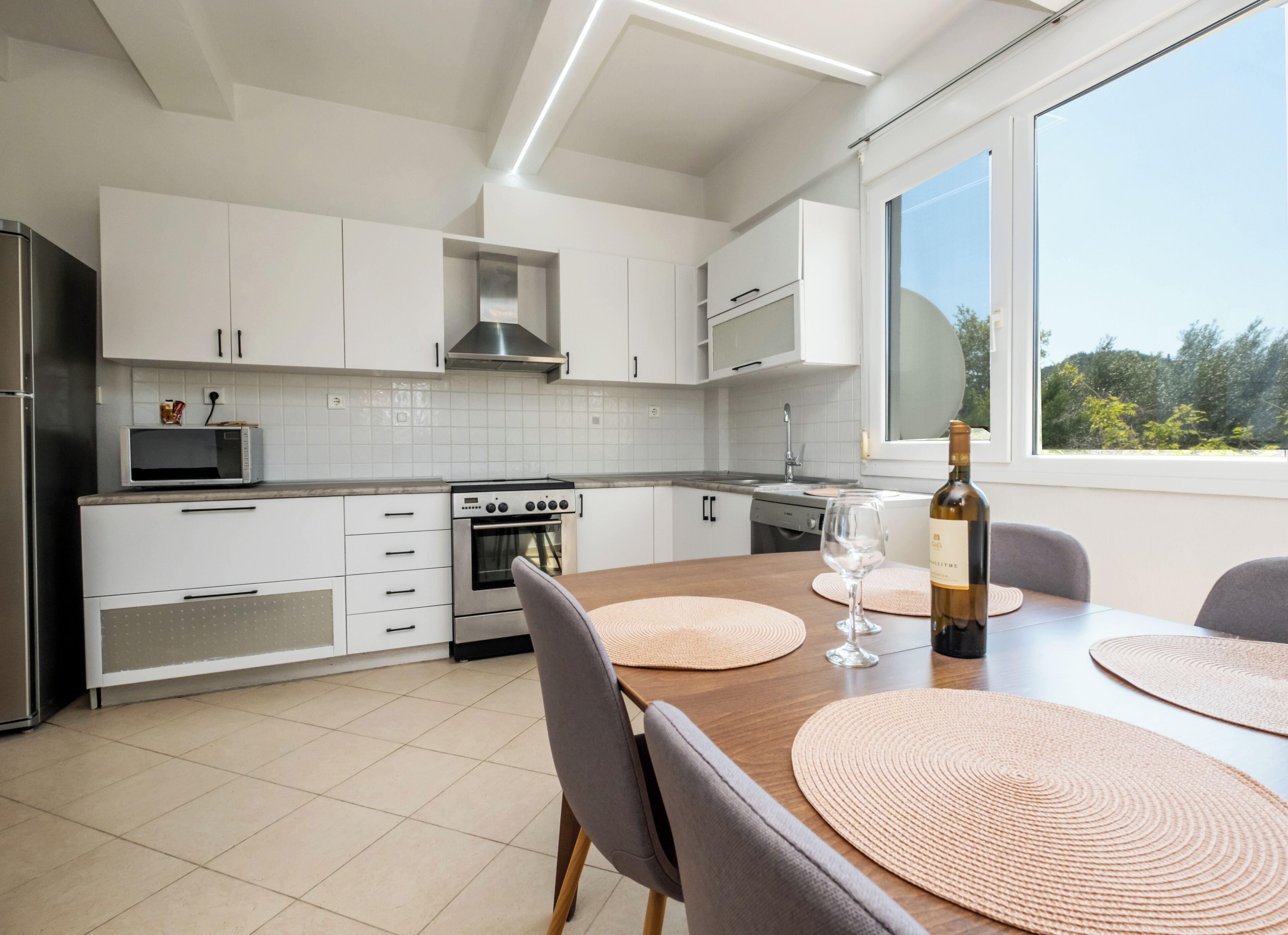 A modern kitchen with white cabinets, a built-in oven, and a dining table with placemats and a wine bottle.
