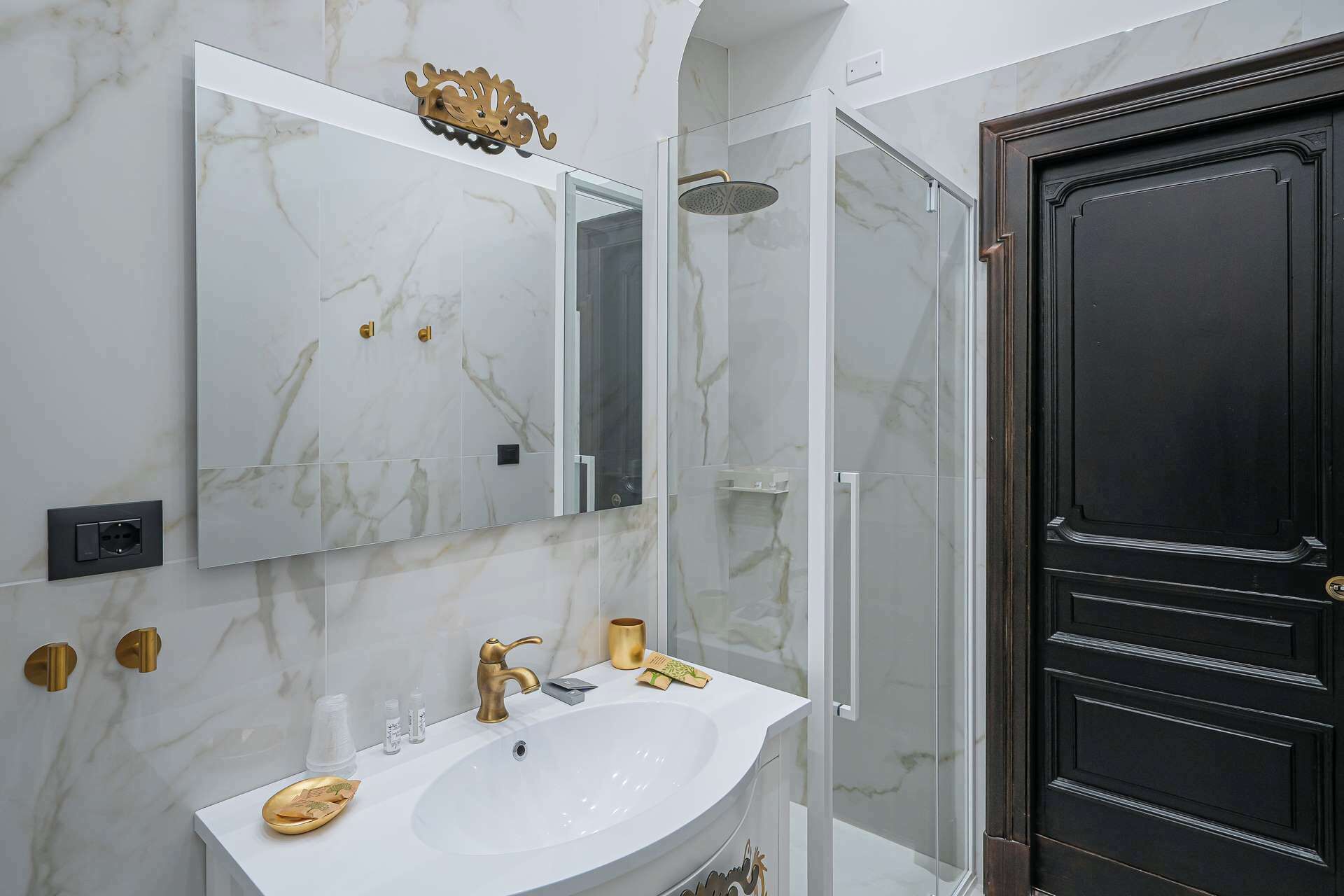 A bathroom with a white sink, gold fixtures, a glass shower enclosure, and a dark wooden door.