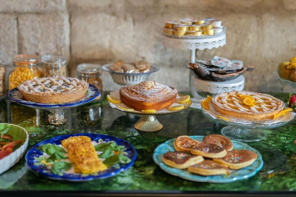 Diversos postres a la vista, como pasteles y hojarascas, sobre una mesa de vidrio frente a una pared de piedra.