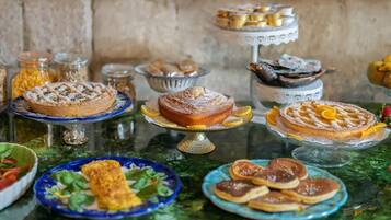 Diversos postres a la vista, como pasteles y hojarascas, sobre una mesa de vidrio frente a una pared de piedra.